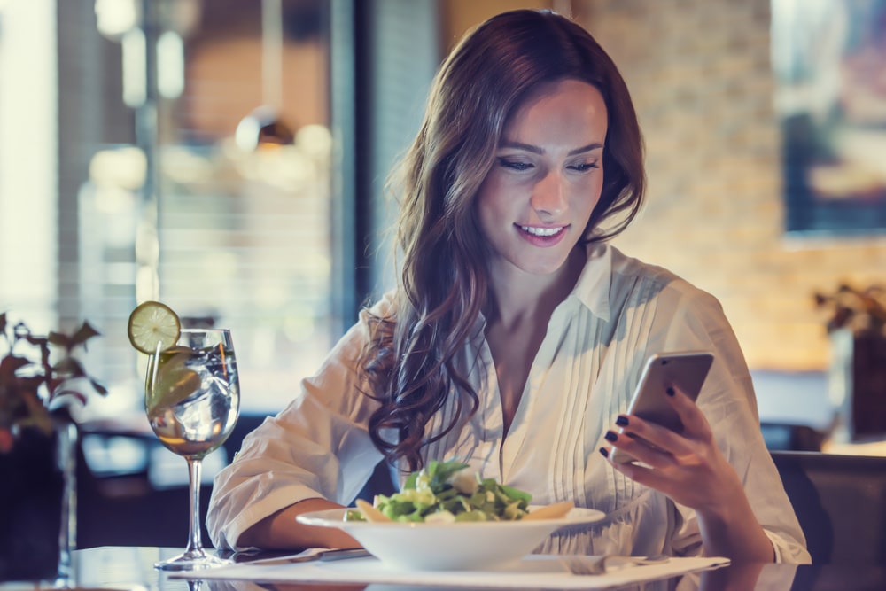 Eine schöne junge Frau verwendet eine Anwendung, um eine SMS-Nachricht auf ihrem Smartphone zu senden, während sie im Restaurant einen Salat isst.