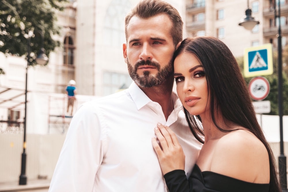 A stylish couple is captured in the streets of Europe, striking poses for the camera. The woman exudes elegance in her black evening dress, while her dapper boyfriend complements her look with his sharp suit. The man's rugged charm adds to the overall allure of the scene, as they both enjoy each other's company outdoors.