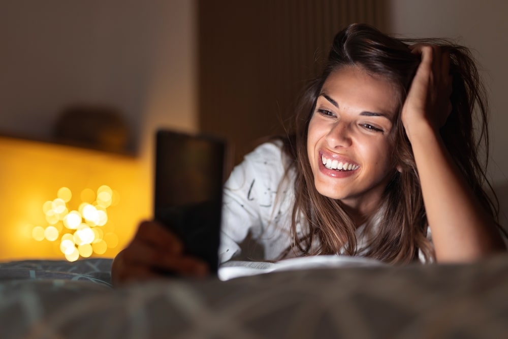 Fröhliche junge Frau liegt nachts im Bett und führt einen Videoanruf mit einem Smartphone