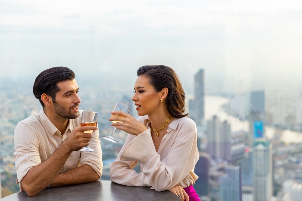 Ein kaukasisches Paar feiert fröhlich auf einer luxuriösen Dachterrassenbar eines Wolkenkratzers während eines Sommer-Sonnuntergangs. Der selbstbewusste Mann und die Frau genießen den Stadtlebensstil und haben Abendessen sowie Cocktails in einem Außenrestaurant