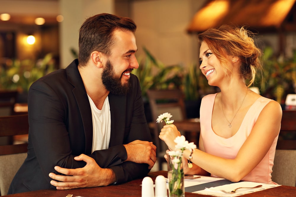 Ein junges Paar sitzt in einem Restaurant, während die Frau in einem festlichen Kleid eine Blume hält