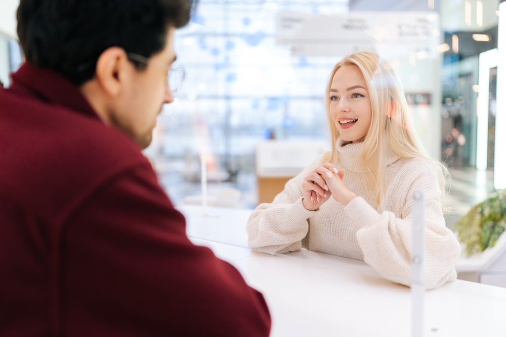 View from behind a man wearing glasses engaged in a conversation with a blonde woman standing behind a glass partition at a well-lit area within a shopping mall. This scenario illustrates the concepts of social distancing and professional interaction in a public space