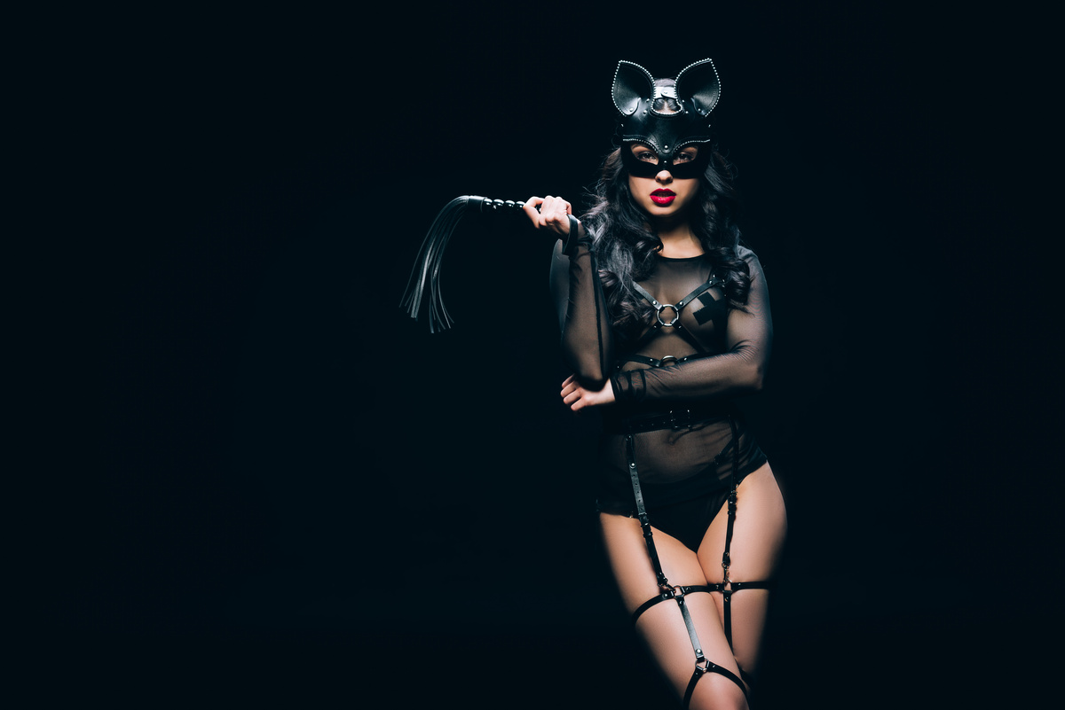 sexy junge brünette Frau in BDSM-Kostüm und Maske mit Leder Auspeitschung Peitsche isoliert auf schwarz