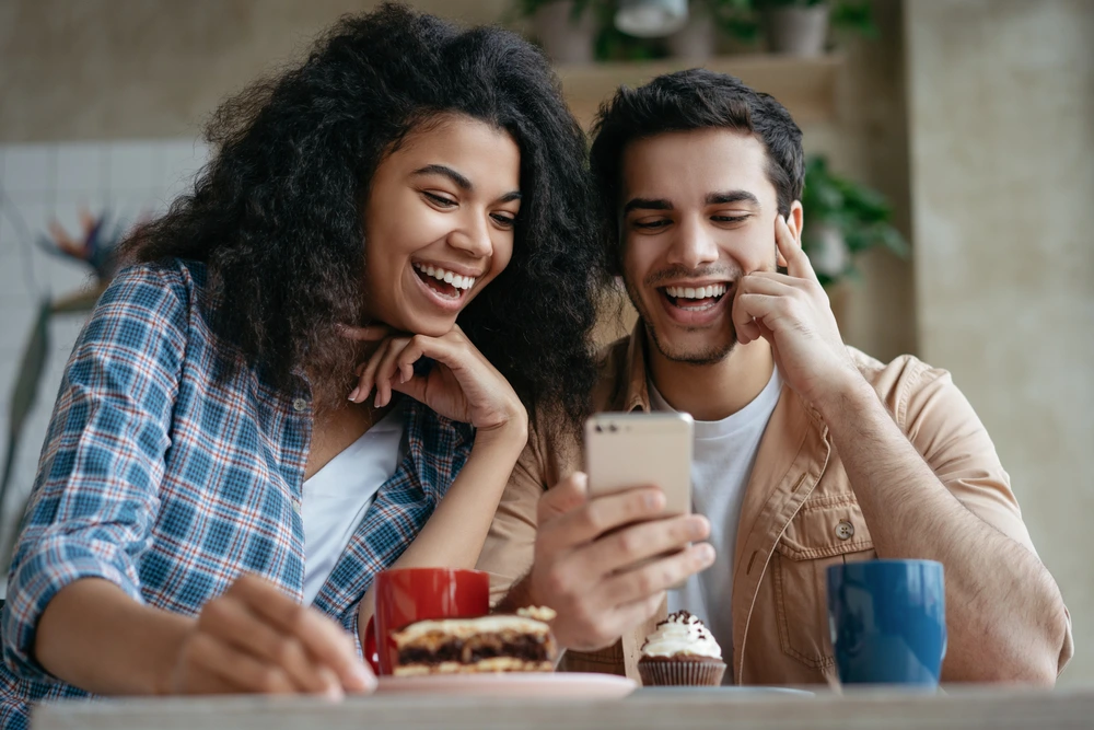 Junges Paar lacht im Café und schaut gemeinsam auf ein Smartphone, Symbol für Spaß und Nähe.