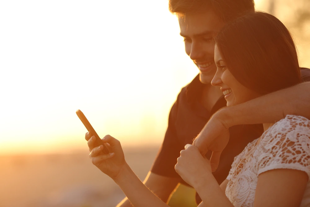 Lächelndes Paar schaut gemeinsam auf ein Smartphone im Sonnenuntergang